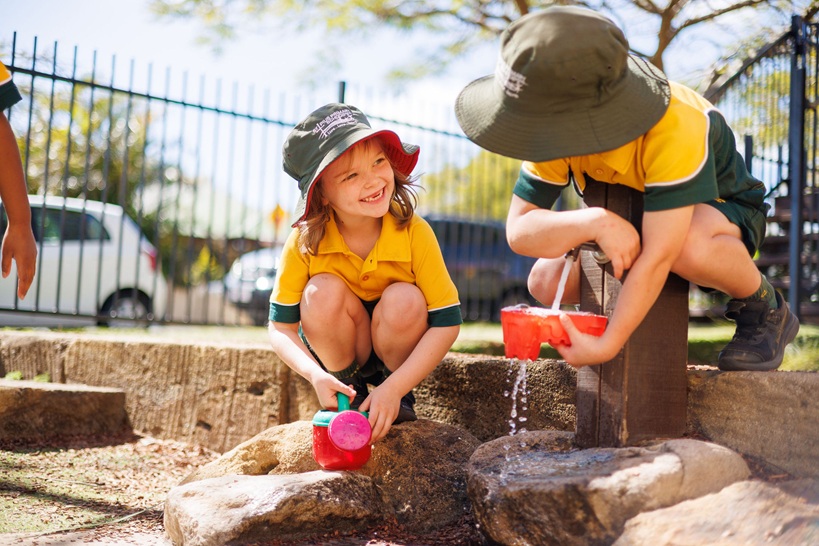 Children in school uniforms playing with water using containers at an outdoor hand pump
