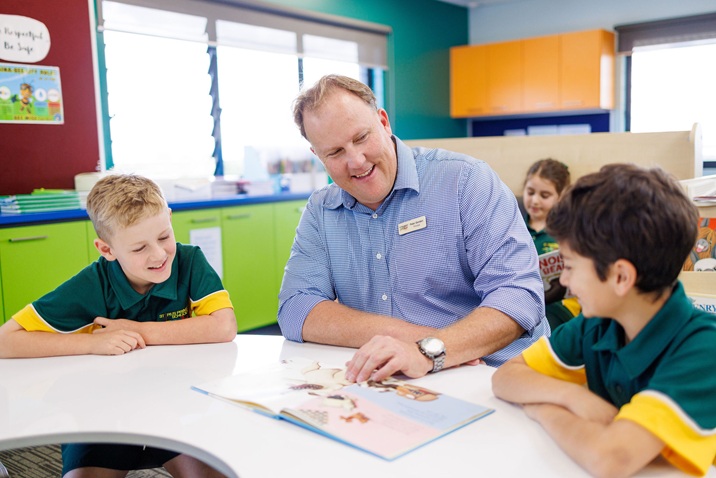 The principle reading a book with two students seated at a classroom table