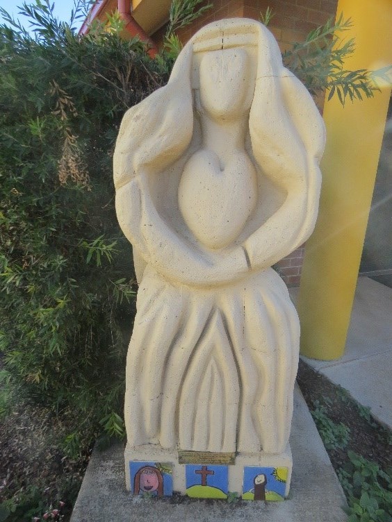 Stone statue of a robed figure holding a heart, featuring small decorative tiles with religious symbols at the base